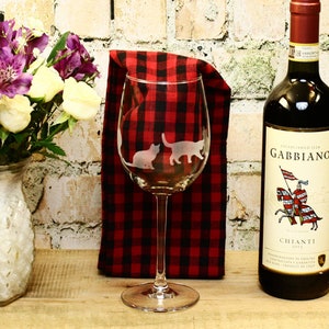 May include: A clear wine glass with two etched cats on it. The glass is sitting on a red and black checkered cloth. A bottle of red wine is in the background.