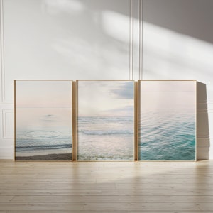Könnte beinhalten: Drei gerahmte Fotos einer Meereslandschaft mit blauem Wasser und einem hellrosa Himmel. Die Fotos sind in einer Reihe auf einem hellen Holzboden angeordnet.