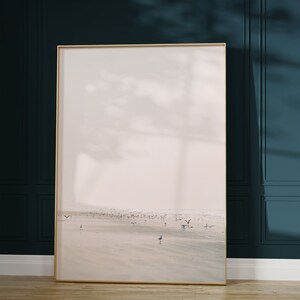 May include: A framed landscape art print featuring a beach scene with birds in flight and on the sand. The artwork has a gold frame and is displayed against a dark teal wall. The floor is light brown wood.