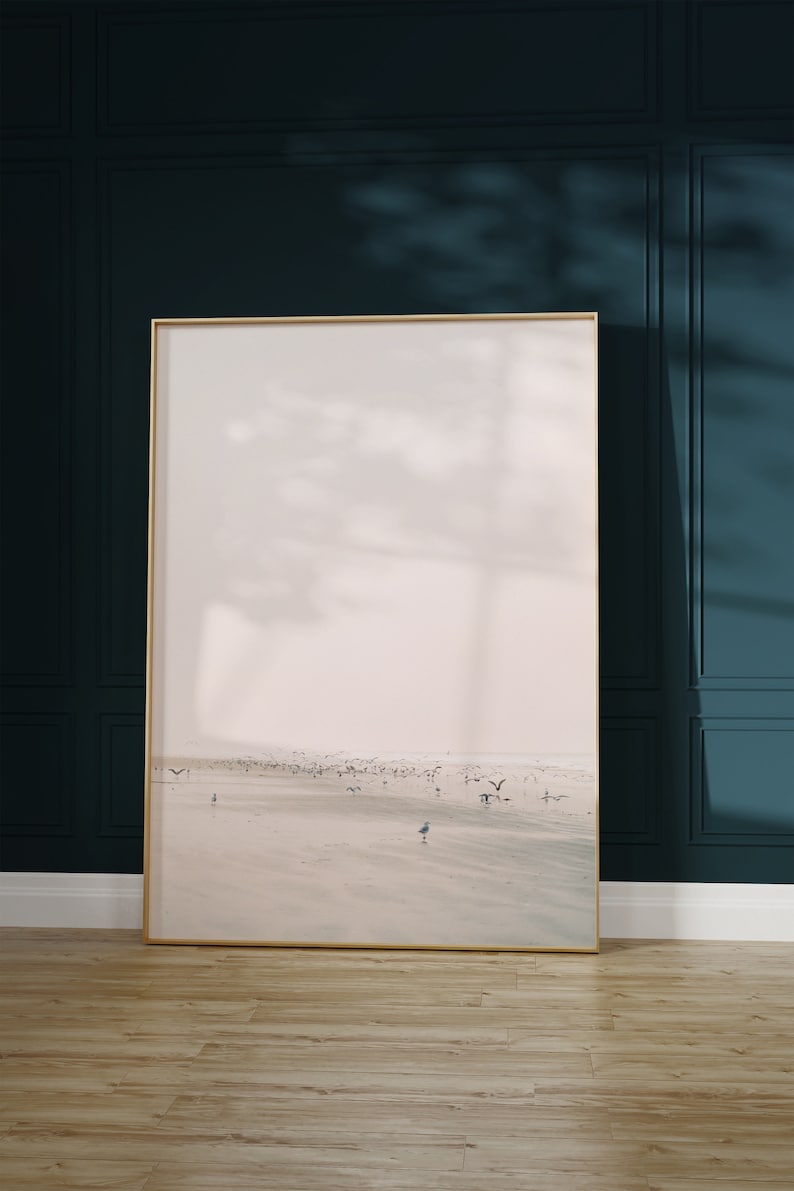 May include: A framed landscape art print featuring a beach scene with birds in flight and on the sand. The artwork has a gold frame and is displayed against a dark teal wall. The floor is light brown wood.