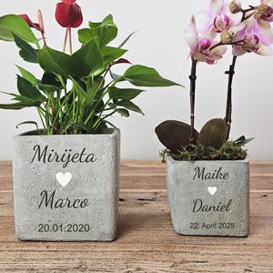 Könnte beinhalten: Zwei quadratische, graue Betonpflanzgefäße mit Pflanzen. Eines hat eine rote Blume und grüne Blätter, das andere eine rosa Orchidee. Jeder Pflanztopf hat einen Namen, ein Herz und ein Datum aufgedruckt.