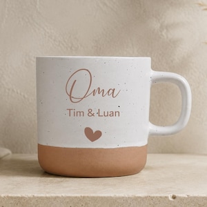 Könnte beinhalten: Ein weißer Keramikbecher mit gesprenkelter Oberfläche und einem terrakottafarbenen Boden. Auf der Vorderseite befinden sich der Schriftzug "Oma" in Schreibschrift, "Tim & Luan" in serifenloser Schrift und ein kleines Herz in Braun.