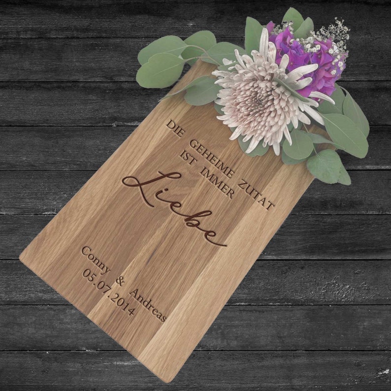 Op de afbeelding: Een houten snijplank met de tekst "Die geheime Zutat ist immer Liebe" en "Conny & Andreas 05.07.2014" erop gegraveerd. De plank is versierd met een boeket witte en paarse bloemen.