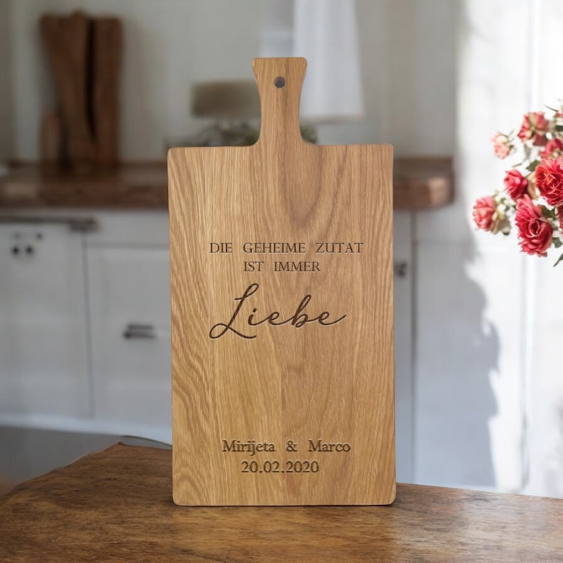 Op de afbeelding: Een houten snijplank met de tekst "Die geheime Zutat ist immer Liebe" en "Mirjjeta & Marco 20.02.2020" erop gegraveerd.