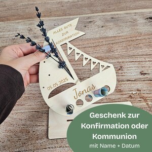 K&ouml;nnte beinhalten: Ein Geschenkhalter in Form eines Holzsegelschiffs mit dem Namen "Jonas" und dem Datum "05.05.2025" eingraviert. Das Segelboot hat ein Banner mit der Aufschrift "ALLES GUTE ZUR KONFIRMATION". Es ist mit einem kleinen Bouquet und einem gr&uuml;nen Oval mit Text verziert.