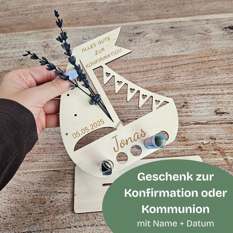 K&ouml;nnte beinhalten: Ein Geschenkhalter in Form eines Holzsegelschiffs mit dem Namen "Jonas" und dem Datum "05.05.2025" eingraviert. Das Segelboot hat ein Banner mit der Aufschrift "ALLES GUTE ZUR KONFIRMATION". Es ist mit einem kleinen Bouquet und einem gr&uuml;nen Oval mit Text verziert.