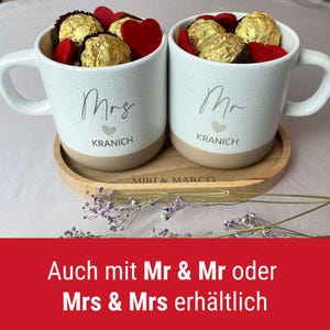 May include: Two white speckled mugs with the words "Mrs" and "Mr" printed on them, filled with gold-wrapped chocolates and red heart-shaped decorations. The mugs sit on a wooden tray with the names "KRANICH" and "MIRI & MARCO" printed on them.