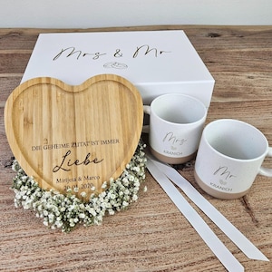 Könnte beinhalten: Ein herzförmiges Bambustablett mit der Gravur DIE GEHEIME ZUTAT IST IMMER Liebe Mirijeta & Marco 20.01.2020, zwei gesprenkelte Keramiktassen mit der Aufschrift Mrs Kranich und Mr Kranich sowie eine weiße Geschenkbox mit Mrs & Mr und Ring-Symbolen.