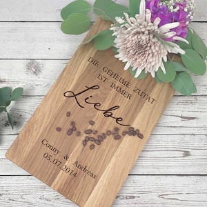 Op de afbeelding: Een houten snijplank met de tekst "Die geheime Zutat ist immer Liebe" en "Conny & Andreas 05.07.2014" erop gegraveerd. De plank is versierd met koffiebonen en een boeket bloemen.