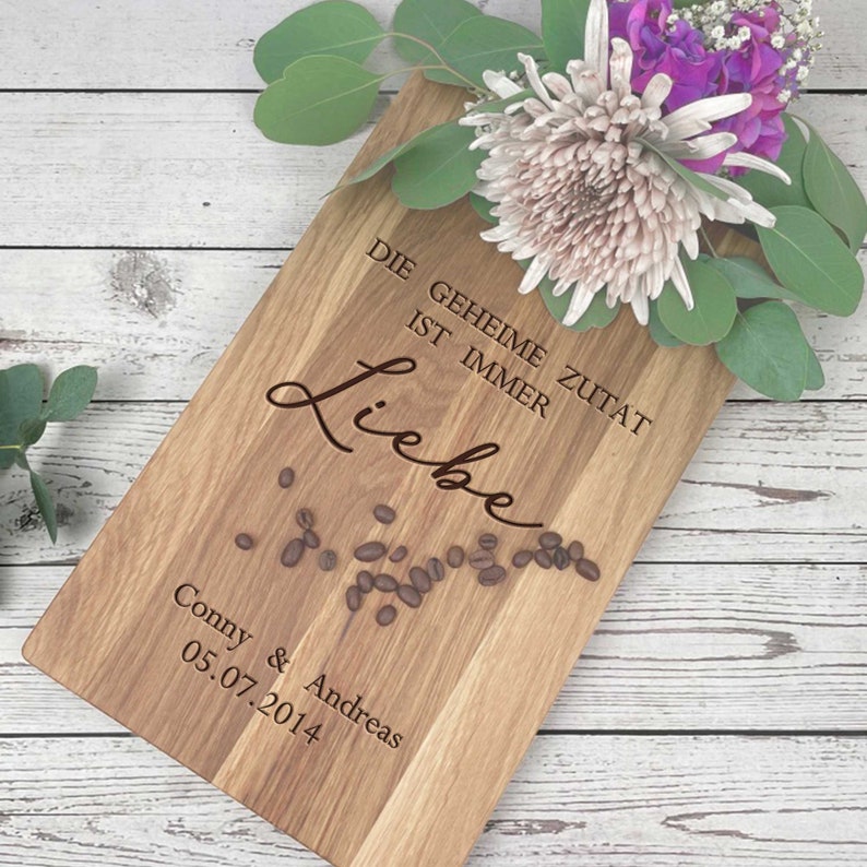 Op de afbeelding: Een houten snijplank met de tekst "Die geheime Zutat ist immer Liebe" en "Conny & Andreas 05.07.2014" erop gegraveerd. De plank is versierd met koffiebonen en een boeket bloemen.