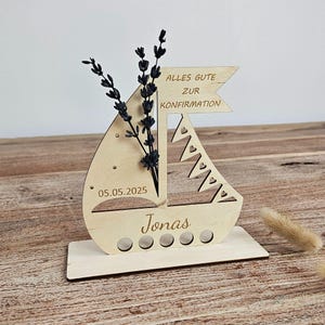 K&ouml;nnte beinhalten: Ein Holzdekor in Form eines Segelboots mit dem Text "Alles Gute zur Konfirmation" und dem Namen "Jonas". Das Datum "05.05.2025" ist eingraviert. Das Segelboot enth&auml;lt getrocknete Blumen.