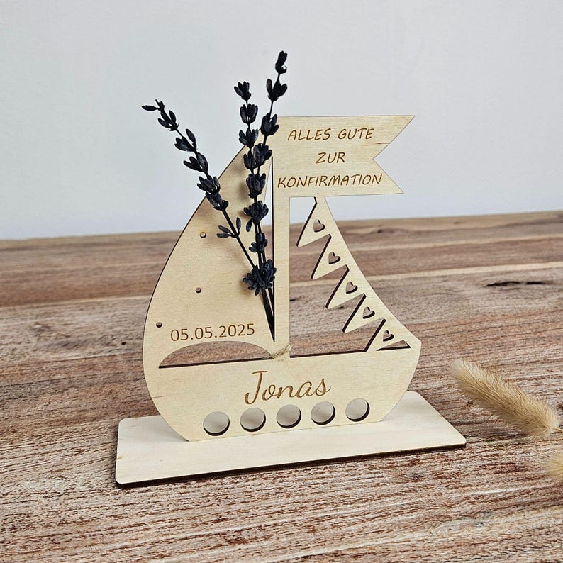 K&ouml;nnte beinhalten: Ein Holzdekor in Form eines Segelboots mit dem Text "Alles Gute zur Konfirmation" und dem Namen "Jonas". Das Datum "05.05.2025" ist eingraviert. Das Segelboot enth&auml;lt getrocknete Blumen.