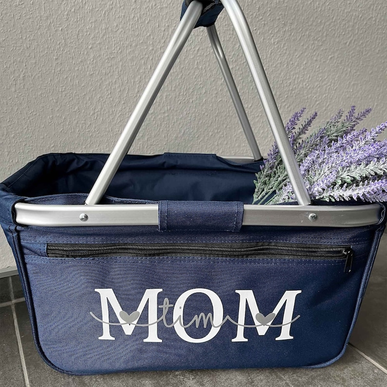 May include: A navy blue canvas tote bag with a silver metal frame and handles. The bag has a zippered pocket and the word "MOM" written in white with two small hearts on either side of the word.