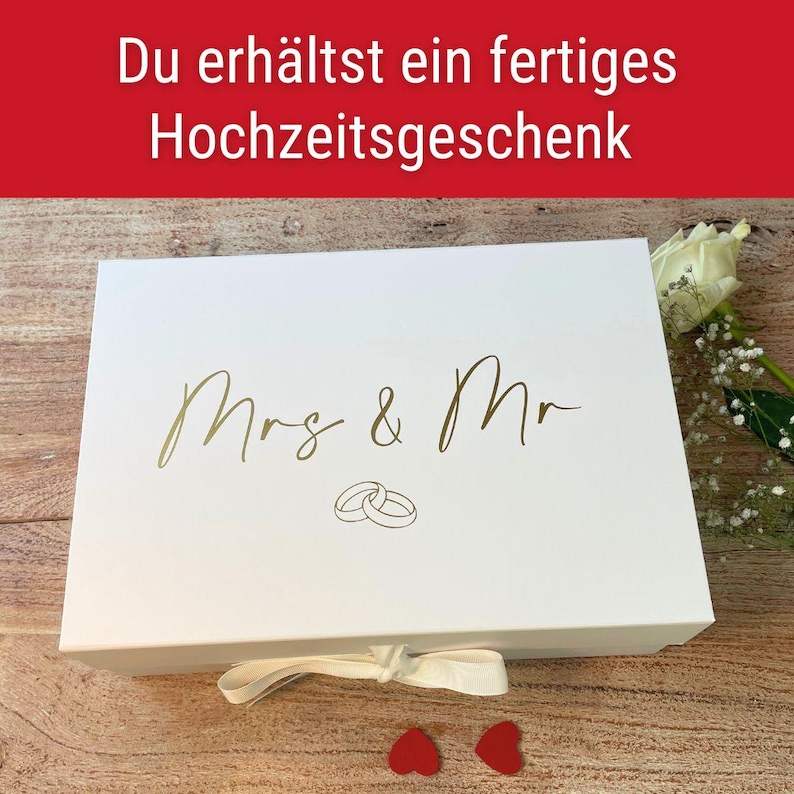 May include: A white gift box with gold lettering that reads "Mrs & Mr" and two interlocking rings. The box has a white ribbon and two red heart-shaped decorations. The text at the top says "You get a finished wedding gift."