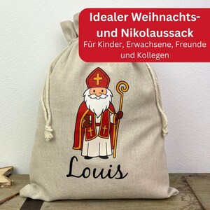 May include: A beige drawstring gift bag featuring a cartoon illustration of Saint Nicholas in red and white robes, holding a staff. The name "Louis" is printed in black script below the illustration. Text in a red banner reads "Ideal Christmas and St. Nicholas Sack."