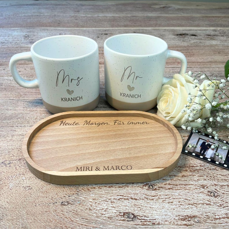 May include: A set of two white ceramic mugs with the words "Mrs" and "Mr" printed on them, along with a wooden tray. The mugs have a speckled design and a tan base. The tray has the text "Heute. Morgen. F&uuml;r immer" and "MIRI & MARCO".