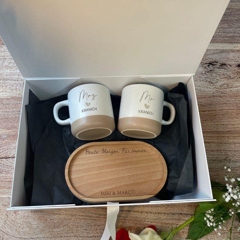 May include: A gift set in a white box containing two white and tan mugs with "Mrs" and "Mr" printed on them, along with a wooden serving tray. The mugs have the name "KRANICH" printed below the titles. The tray is engraved with "Heute Morgen F&uuml;r immer" and "MIRI & MARCO".