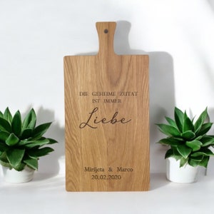 Op de afbeelding: Een houten snijplank met een handvat, gegraveerd met de tekst "Die geheime Zutat ist immer Liebe" en "Mirjeta & Marco 20.02.2020".