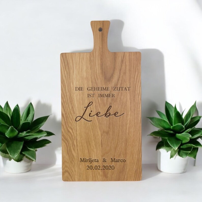 Op de afbeelding: Een houten snijplank met een handvat, gegraveerd met de tekst "Die geheime Zutat ist immer Liebe" en "Mirjeta & Marco 20.02.2020".