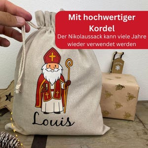 May include: A beige drawstring gift bag featuring a cartoon illustration of Santa Claus in red and white robes, holding a staff. The name "Louis" is printed in black script. A wrapped gift box is in the background.