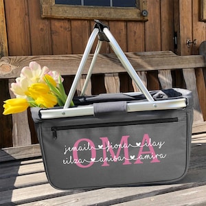 May include: Gray canvas tote bag with a zipper closure and two silver handles. The bag has pink lettering that says "OMA" with the names "jamail, fiona, ilay, eliza, anisa, amira" written underneath in a smaller font.