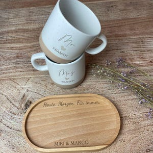 May include: Two white speckled ceramic mugs, one stacked on top of the other, with the words "Mr" and "Mrs" in script. The mugs have a tan base and the name "Kranich" is printed below. A wooden serving tray with the text "Heute. Morgen. F&uuml;r immer." and "MIRI & MARCO" is in the foreground.