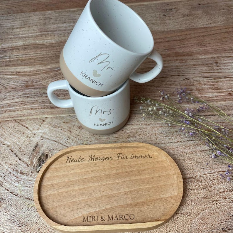 May include: Two white speckled ceramic mugs, one stacked on top of the other, with the words "Mr" and "Mrs" in script. The mugs have a tan base and the name "Kranich" is printed below. A wooden serving tray with the text "Heute. Morgen. F&uuml;r immer." and "MIRI & MARCO" is in the foreground.
