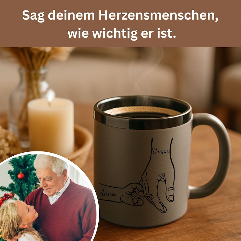 Op de afbeelding: Een grijze koffiemok met een zwarte rand en handvat, gevuld met koffie. De mok heeft een lijntekening van twee handen die elkaar vasthouden, met de namen "Anna" en "Uropa" erop geschreven. De tekst boven de mok luidt "Zeg tegen je geliefden hoe belangrijk ze zijn."