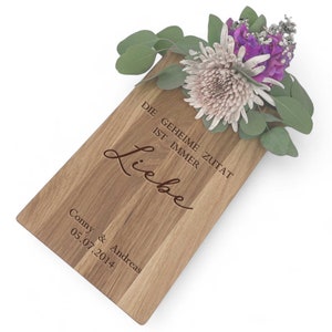 Op de afbeelding: Een houten snijplank met de tekst "Die geheime Zutat ist immer Liebe" en "Conny & Andreas 05.07.2014" erop gegraveerd. De plank heeft een bloemstuk in een vaas.