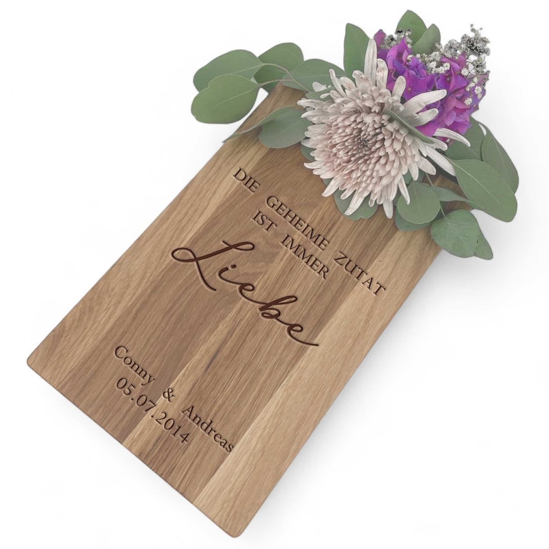 Op de afbeelding: Een houten snijplank met de tekst "Die geheime Zutat ist immer Liebe" en "Conny & Andreas 05.07.2014" erop gegraveerd. De plank heeft een bloemstuk in een vaas.