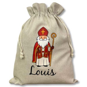 May include: A beige drawstring gift bag featuring a cartoon illustration of a saint in red and white robes, holding a staff. The name "Louis" is printed in black script below the illustration. The bag is made of a natural-looking fabric.