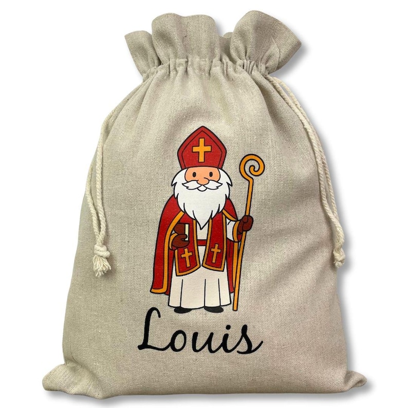 May include: A beige drawstring gift bag featuring a cartoon illustration of a saint in red and white robes, holding a staff. The name "Louis" is printed in black script below the illustration. The bag is made of a natural-looking fabric.