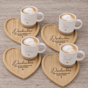 Könnte beinhalten: Vier Keramiktassen mit schaumgekröntem Kaffee, jede personalisiert mit einem Namen und einem kleinen Herz. Die Tassen stehen auf vier herzförmigen Holzuntersetzern, die mit dem Text Dankeschön für all die grossen und kleinen Dinge graviert sind.