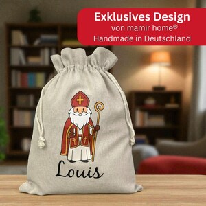 May include: A beige drawstring gift bag featuring a cartoon illustration of a saint in red and white robes, holding a staff and a book. The name "Louis" is written in cursive below the illustration. The bag has the text "Exklusives Design" and "Handmade in Deutschland".