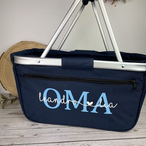 May include: A navy blue canvas tote bag with a zippered pocket and a silver metal frame. The bag has the text "OMA" in light blue with a small white heart and the text "leandro" in white.