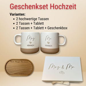 May include: A wedding gift set featuring two white mugs with "Mrs" and "Mr" printed on them, along with a wooden tray and a gift box. The mugs have a speckled design and a tan base. The tray is engraved with text. The gift box is white with gold lettering.