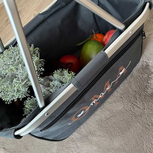 May include: A gray canvas shopping basket with a silver frame and a zipper closure. The basket is filled with fruit and greenery. The word "OMA" is embroidered on the side of the basket in brown cursive.