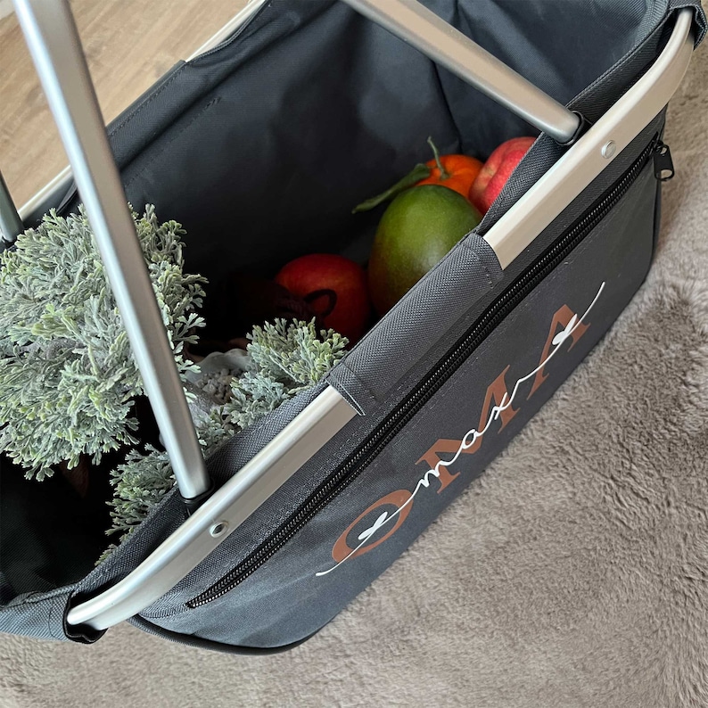 May include: A gray canvas shopping basket with a silver frame and a zipper closure. The basket is filled with fruit and greenery. The word "OMA" is embroidered on the side of the basket in brown cursive.
