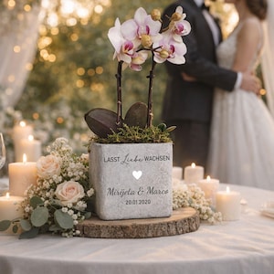 Può includere: Una fioriera quadrata in cemento grigio con un'orchidea, muschio e il testo "LASST Liebe WACHSEN" e nomi. La fioriera è posizionata su una fetta di legno, circondata da candele e fiori, creando un centrotavola romantico per matrimoni.