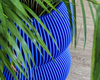 May include: A vibrant blue, ribbed planter pot holds a lush green plant with long, slender leaves. The pot has a unique, textured design with vertical grooves. The plant and pot sit on a light-colored wooden surface.