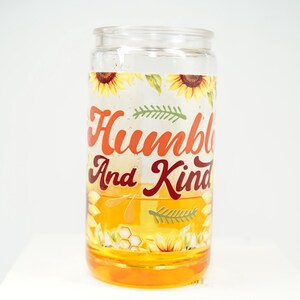 May include: A clear glass jar with a yellow liquid inside. The jar has a decorative label with sunflowers, green leaves, and the words "Humble And Kind" in orange and burgundy lettering. The jar is on a white surface.