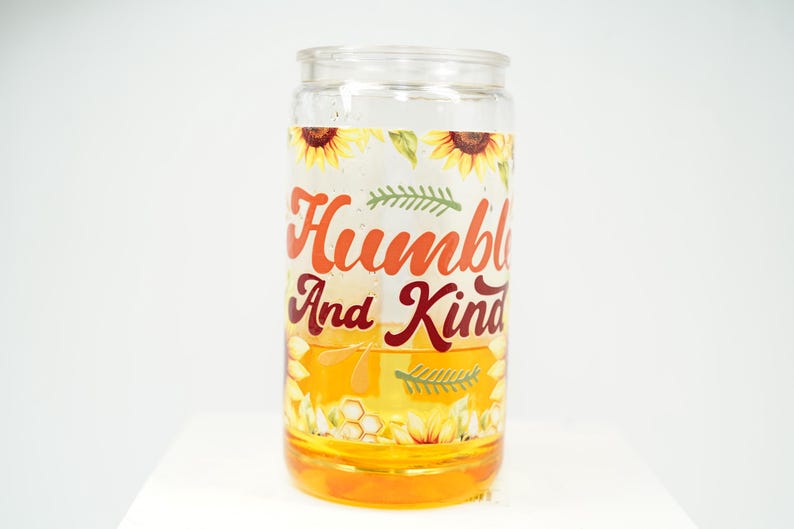 May include: A clear glass jar with a yellow liquid inside. The jar has a decorative label with sunflowers, green leaves, and the words "Humble And Kind" in orange and burgundy lettering. The jar is on a white surface.