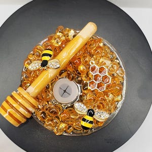 May include: A decorative candle holder with a bee theme. The holder is filled with amber-colored glass beads and features a wooden honey dipper, two bee charms with rhinestone wings, and a honeycomb charm. The holder sits on a black surface.