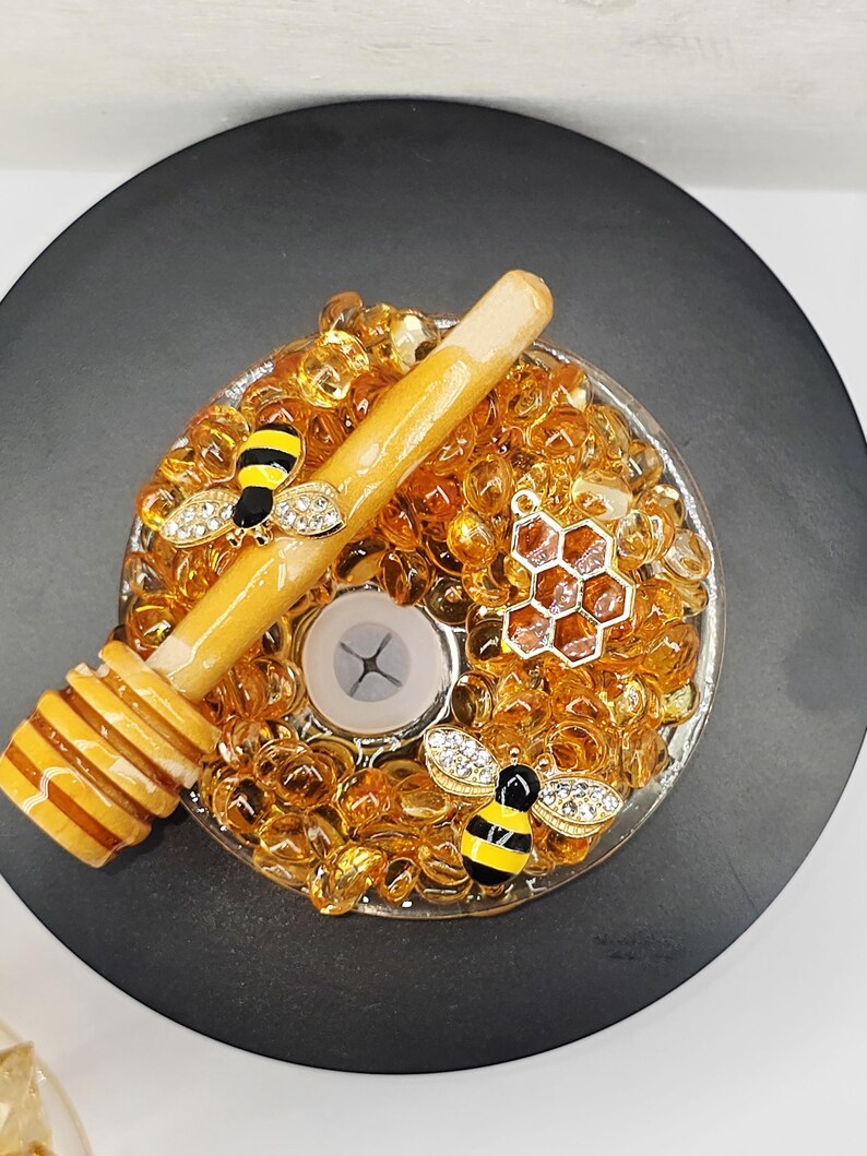 May include: A decorative candle holder with a bee theme. The holder is filled with amber-colored glass beads and features a wooden honey dipper, two bee charms with rhinestone wings, and a honeycomb charm. The holder sits on a black surface.