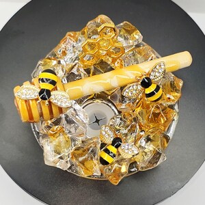 May include: A decorative bottle stopper featuring a bee theme. The stopper has a gold honeycomb design, a honey dipper, and three bee embellishments with crystal accents. The stopper is set on a black base.