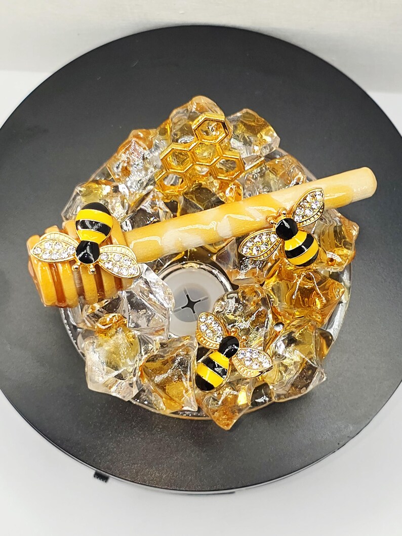 May include: A decorative bottle stopper featuring a bee theme. The stopper has a gold honeycomb design, a honey dipper, and three bee embellishments with crystal accents. The stopper is set on a black base.