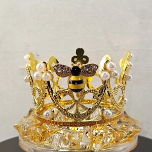 May include: A gold-colored crown with a bee design, adorned with faux pearls. The bee has black and yellow stripes and is embellished with rhinestones. The crown sits atop a clear, faceted base.