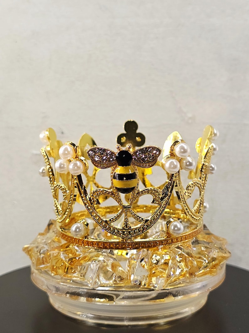 May include: A gold-colored crown with a bee design, adorned with faux pearls. The bee has black and yellow stripes and is embellished with rhinestones. The crown sits atop a clear, faceted base.