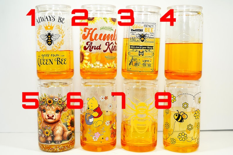 May include: Eight clear glass tumblers with yellow liquid inside. Each glass has a different design, including bees, sunflowers, and cartoon characters. The glasses are filled with a yellow liquid, likely honey or a similar beverage.