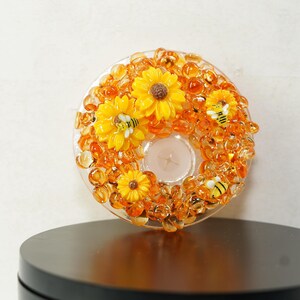 May include: A decorative candle holder with a clear glass base, adorned with yellow sunflowers, orange beads, and small bee figurines. The candle holder is designed to hold a single tea light.
